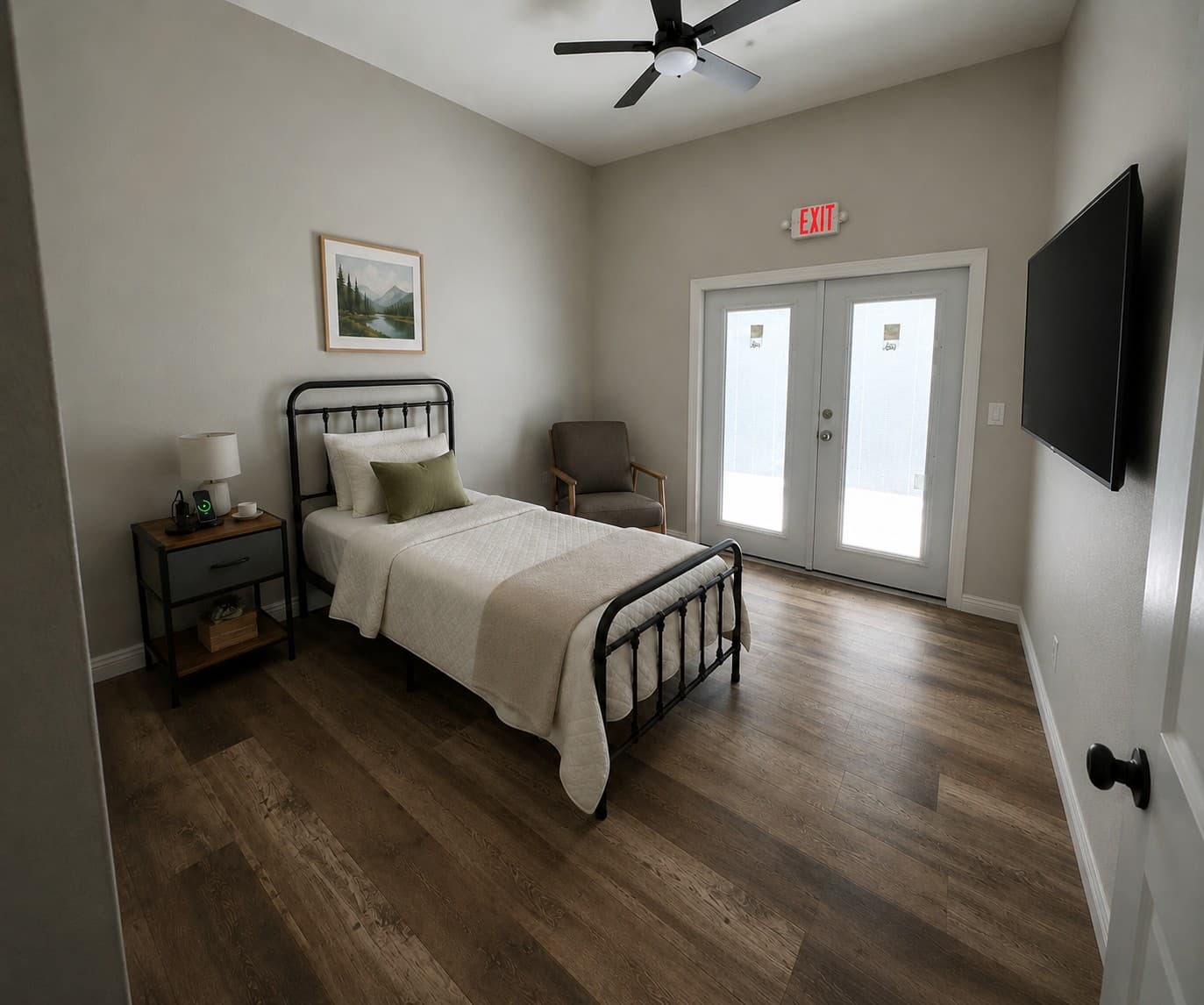 Neutral bedroom with a single black metal bed, armchair, and glass doors with exit sign.